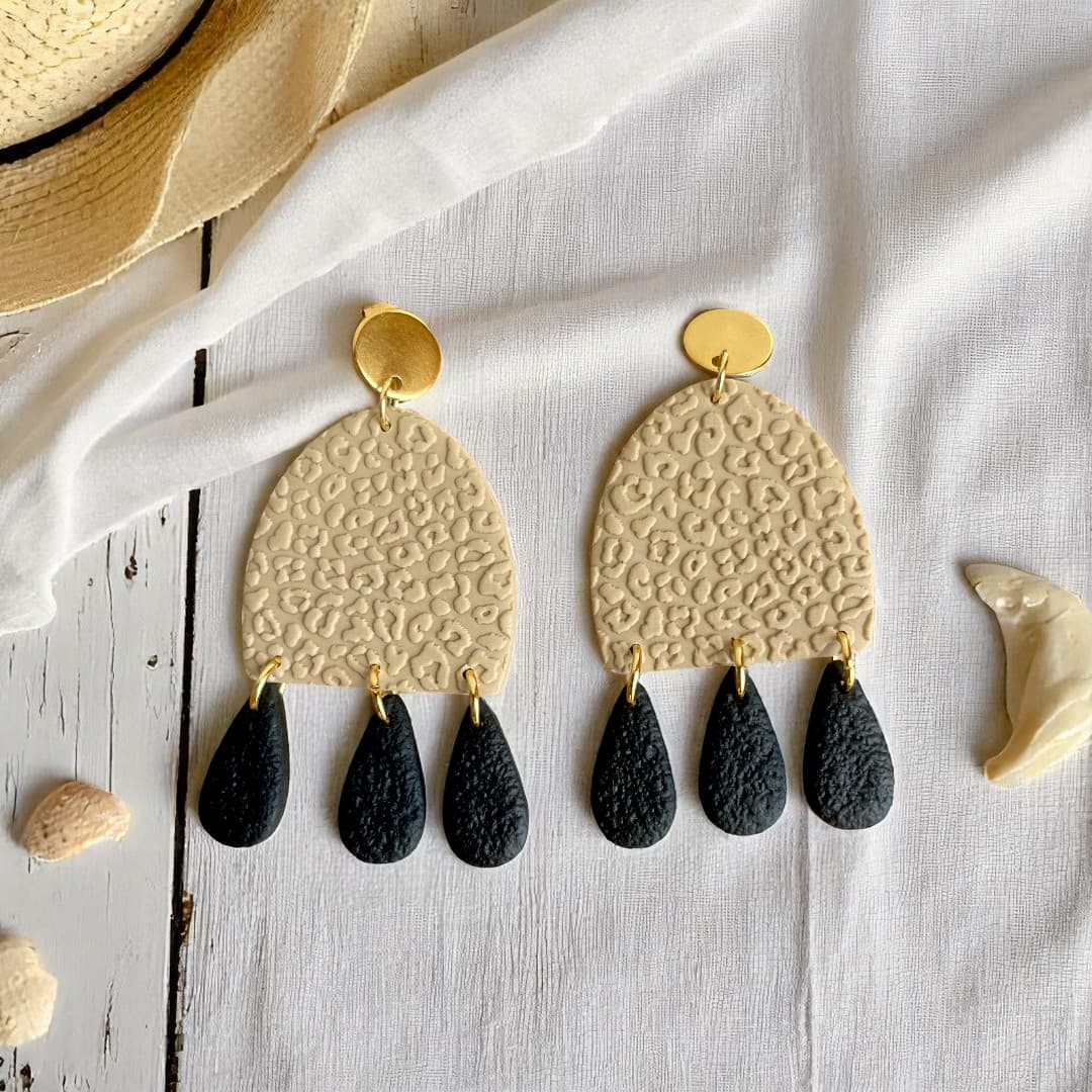 Boucles d’oreilles pendantes en polymère beige avec motif léopard texturé, pampilles noires texturées et attaches dorées.